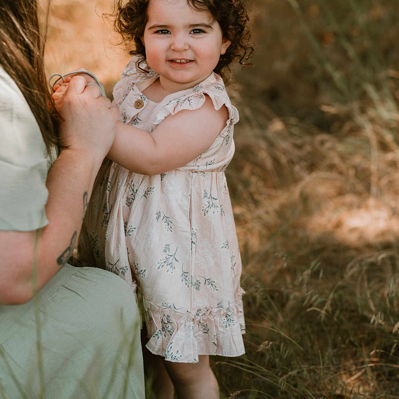 Natalie is registered to the contest to win money with this photo: child, toddler, curly_hair, dress, holding_hands, adult, nature, outdoor, grass, flowers, smile, happy, person, portrait, sunlight, summer, field, family, casual, playful