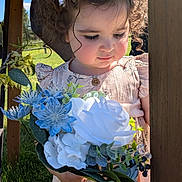Natalie is registered to the contest to win money with this photo: child, toddler, curly_hair, bow, flower_bouquet, blue_flowers, white_flowers, dress, outdoor, sunlight, green_grass, wooden_post, nature, portrait, cute, young_child, person, holding, daylight, garden