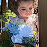 child, toddler, curly_hair, bow, flower_bouquet, blue_flowers, white_flowers, dress, outdoor, sunlight, green_grass, wooden_post, nature, portrait, cute, young_child, person, holding, daylight, garden