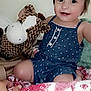 baby, blue_romper, child, cute, face, giraffe, hand, happy, headband, indoors, patterned_fabric, person, plush_toy, quilt, red_and_white, sitting, smiling, soft_toy, toddler, white_bow