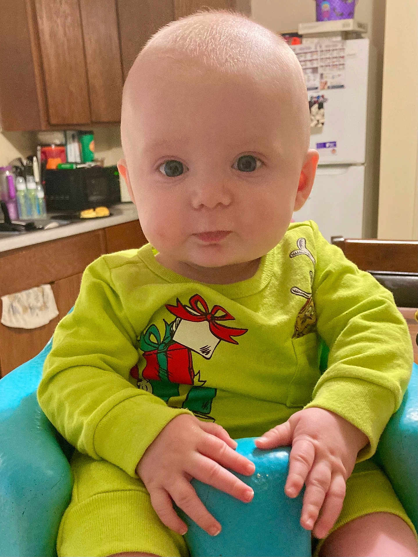 Cayson is registered to the contest to win money with this photo: baby, child, infant, green_onesie, blue_baby_chair, kitchen, cabinet, refrigerator, microwave, countertop, hands, face, eyes, ears, table, furniture, portrait, home, indoor, curious