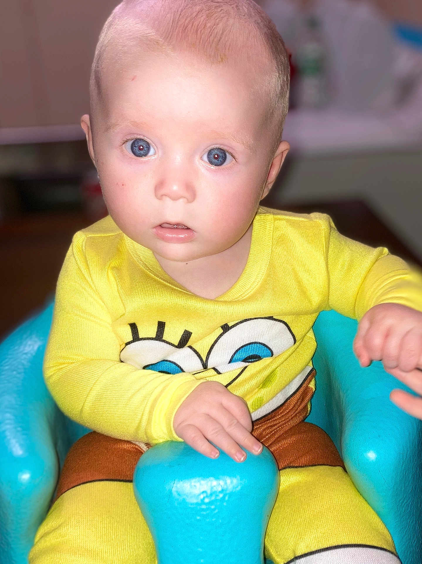 Cayson is registered to the contest to win money with this photo: baby, child, infant, yellow_clothing, spongebob, blue_seat, indoor, cute, portrait, face, hand, looking_at_camera, young_child, seated, curious, expression, bright_colors, soft_skin, clothing, person