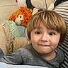 Liam is registered to the contest to win money with this photo: child, boy, toddler, face, smiling, portrait, indoors, bedroom, bed, blanket, stuffed_animal, plush_toy, cow_toy, lion_toy, pillow, bedding, gray_shirt, brown_hair, sitting, phone_screenshot