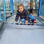 Liam is registered to the contest to win money with this photo: toddler, child, playground, slide, outdoor, park, sitting, smiling, portrait, jacket, winter_clothing, jeans, sneakers, playground_equipment, stroller, tree, buildings, urban, daylight, playful