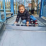 toddler, child, playground, slide, outdoor, park, sitting, smiling, portrait, jacket, winter_clothing, jeans, sneakers, playground_equipment, stroller, tree, buildings, urban, daylight, playful