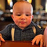 baby, child, face, portrait, indoor, table, clothing, suspenders, blue_shirt, hands, expression, young_child, cute, person, head, skin, cheeks, short_hair, focus, warm_lighting