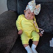 Nia is registered to the contest to win money with this photo: baby, child, yellow_outfit, white_socks, floral_bow, pacifier_clip, strawberry, couch, gray_texture, indoor, cute, infant, smiling, seated, cozy, portrait, person, young_child, happy, comfortable