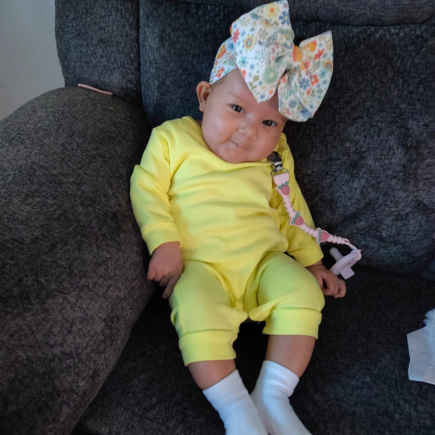Nia is registered to the contest to win money with this photo: baby, child, yellow_outfit, white_socks, floral_bow, pacifier_clip, strawberry, couch, gray_texture, indoor, cute, infant, smiling, seated, cozy, portrait, person, young_child, happy, comfortable
