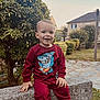 toddler, child, smiling, red_sweatshirt, shark_graphic, bench, stone_bench, garden, bush, tree, house, pathway, grass, outdoors, portrait, hands, sneakers, daytime, happy, casual_clothing