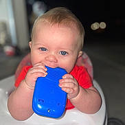 Kaelahni is registered to the contest to win money with this photo: baby, blue_eyes, chewing, child, closeup, face, hand, high_chair, indoor, infant, person, plastic_toy, red_clothing, skin, smiling, toy, young_child, cute, portrait, happy