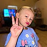 child, boy, peace_sign, puckered_lips, blue_tshirt, minnie_mouse, cartoon_characters, indoor, living_room, blurred_background, television, wooden_floor, casual_clothing, short_hair, portrait, cute, playful, person, gesture, expression