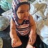baby, child, infant, sitting, striped_hat, bonnet, striped_clothing, onesie, barefoot, feet, hands, face, portrait, indoors, bedroom, bed, blankets, pillows, serious_expression, candid_photo