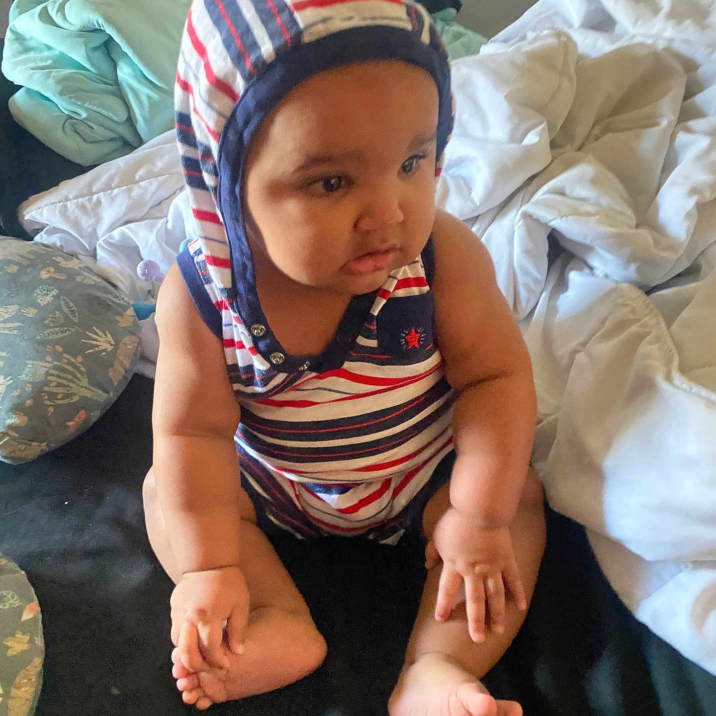 Nathaniel Dorcin joined the competition — help win amazing prizes! baby, child, infant, sitting, striped_hat, bonnet, striped_clothing, onesie, barefoot, feet, hands, face, portrait, indoors, bedroom, bed, blankets, pillows, serious_expression, candid_photo