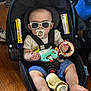 Wade is registered to the contest to win money with this photo: baby, car_seat, sunglasses, pacifier, toy, bottle, infant, child, floor, wooden_floor, blue_shorts, white_shirt, indoor, seat_belt, foot, hand, cute, casual, relaxed, accessory