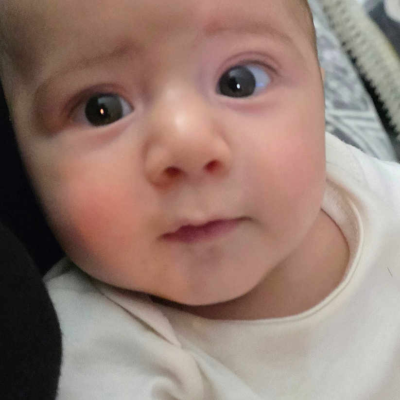 Yasir is registered to the contest to win money with this photo: baby, infant, child, portrait, close_up, face, eyes, wide_eyed, looking_at_camera, indoors, bed, bedding, blanket, clothing, white_outfit, sitting, cozy, home, photography, people