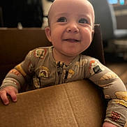 Maison is registered to the contest to win money with this photo: baby, infant, child, smile, cardboard_box, pajamas, hand, portrait, indoor, bokeh, blur, face, eyes, happy, cute, grip, wood_floor, chair, home, soft_lighting