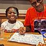 Arianna is registered to the contest to win money with this photo: child, face_paint, smiling, man, glasses, orange_shirt, table, restaurant, menus, crayons, coloring_sheet, plate, condiments, glass, happy, indoor, sign, text, people, dining