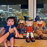 baby, child, infant, scarecrow, dolls, portrait, sitting, held, blue_outfit, yellow_hair, hat, storefront, window, reflection, wooden_bench, indoor, shopping_area, wide_eyed, family_photo, safe