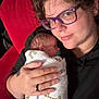 Carter is registered to the contest to win money with this photo: newborn, baby, person, glasses, blanket, giraffe_pattern, couch, red_couch, holding, swaddled, indoor, portrait, closeup, hand, ring, tattoo, black_hoodie, blue_pants, pacifier, curly_hair