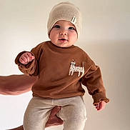 Aylan participe au concours pour gagner de l'argent avec cette photo : baby, child, boy, hat, sweater, pants, hands, face, cute, infant, knit_hat, brown_sweater, cream_pants, indoor, portrait, smiling, holding, expression, light_background, text_overlay