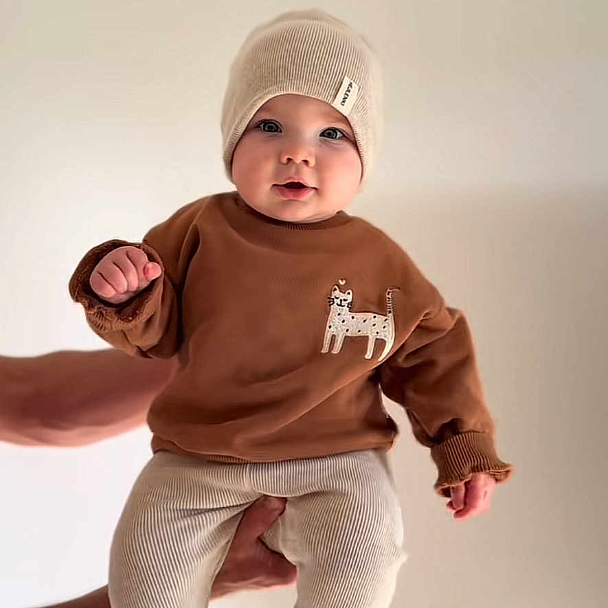 Aylan participe au concours pour gagner de l'argent avec cette photo : baby, child, boy, hat, sweater, pants, hands, face, cute, infant, knit_hat, brown_sweater, cream_pants, indoor, portrait, smiling, holding, expression, light_background, text_overlay