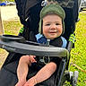 Elijah is registered to the contest to win money with this photo: baby, child, stroller, hat, smiling, outdoor, grass, sunny, happy, young_child, infant, footwear, seat, baby_stroller, person, green, black, daylight, parked, vehicle