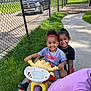 child, children, tricycle, smiling, outdoor, sidewalk, grass, fence, toy, play, happy, person, sunlight, casual_clothing, headband, black_shoes, greenery, park, summer, recreation
