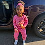 child, girl, pink_clothing, headband, curly_hair, snack, drink, car, car_door, tarmac, sneakers, outdoor, sunlight, standing, casual, young_child, person, daylight, parking_lot, reflection