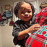 toddler, child, person, indoor, curly_hair, plaid_shirt, adult, hand, bedroom, bags, door, wall, curtain, furniture, clothing, portrait, holding, young_child, warm_lighting, casual