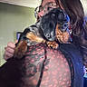 person, dog, puppy, dachshund, glasses, indoor, holding, black_hair, patterned_clothing, mesh_fabric, pet, cute, animal, face, portrait, closeup, warm_lighting, brown_black_coat, looking_away, cuddle