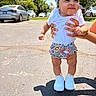 baby, child, smile, white_shirt, floral_headband, floral_shorts, white_sneakers, hands, outdoor, sunny, shadow, pavement, car, trees, person_holding, cute, happy, summer, fashion, portrait