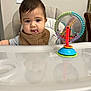 baby, child, high_chair, toy, tongue_out, bib, indoor, face, reflection, hair, playful, colorful, plastic, seat, person, smiling, expression, furniture, light_switch, wooden_chair