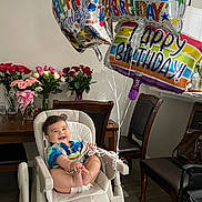 Leon is registered to the contest to win money with this photo: baby, high_chair, balloons, happy_birthday, smile, roses, bouquet, flowers, dining_table, chair, indoor, celebration, diaper, person, wood_floor, colorful, party, joyful, infant, room