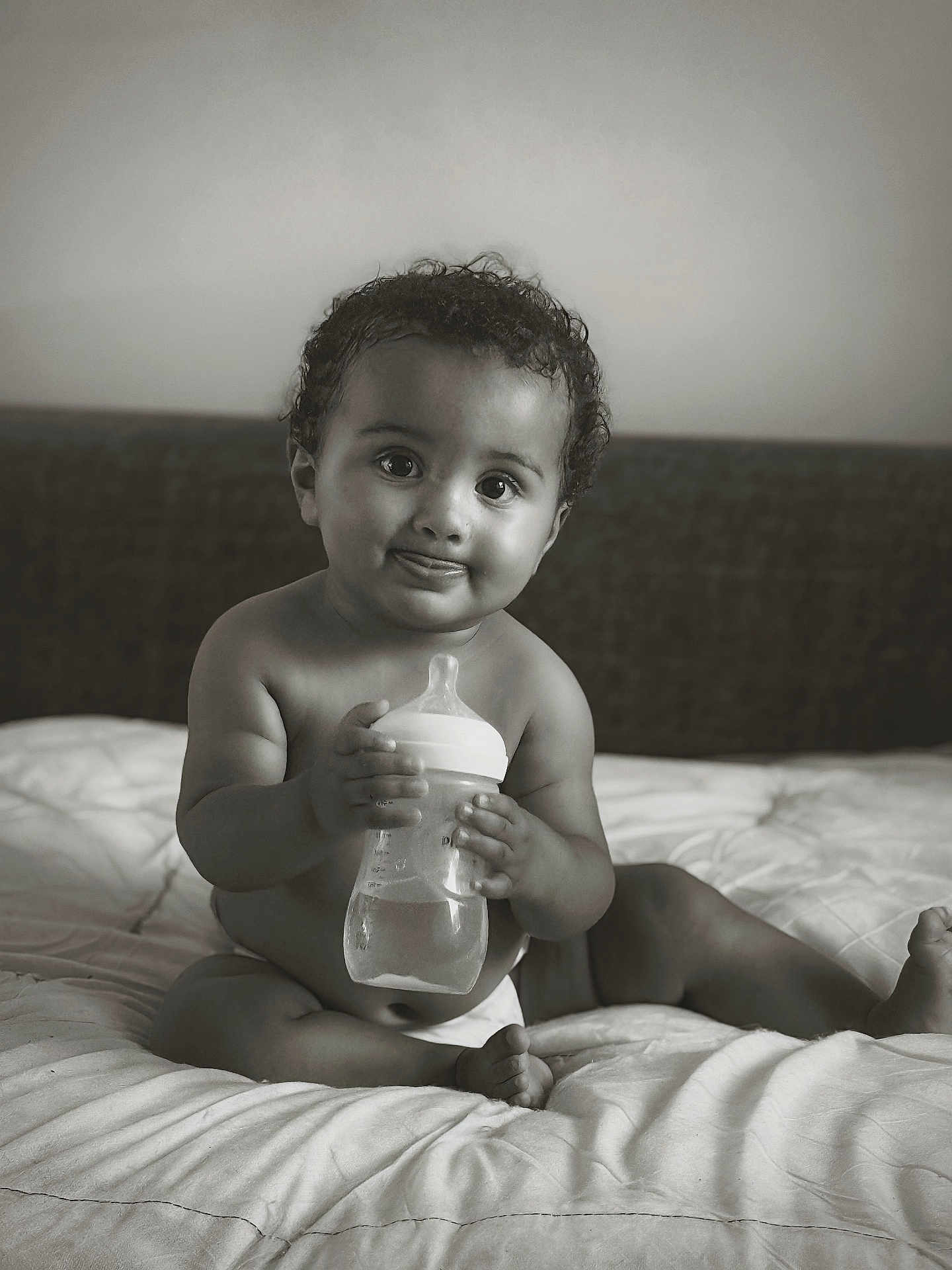 Roy participe au concours pour gagner de l'argent avec cette photo : baby, child, infant, bottle, bed, diaper, black_and_white, curly_hair, cute, smiling, tongue_out, indoors, portrait, sitting, soft_light, expression, person, young, nursery, cozy
