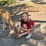 child, girl, deer, animal, feeding, outdoor, sunlight, shadow, smiling, crouching, nature, fence, dirt, casual_clothing, sneakers, happy, young, wildlife, interaction, park