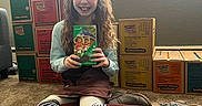 Aubrey is registered to the contest to win money with this photo: boots, brown_skirt, carpet, child, cookie_box, girl, girl_scout_cookies, happy, holding, indoor, long_hair, person, retail, sitting, smile, stacked_boxes, striped_socks, sunlight, window, young