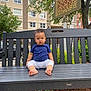 child, toddler, bench, park, outdoor, barefoot, blue_shirt, white_pants, serious_face, sign, tree, building, window, car, urban, sidewalk, grass, daylight, person, nature