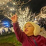 child, toddler, hat, knit_hat, jacket, outdoor, night, lights, trees, festive, holiday_lights, grass, park, people, winter, warm_clothing, reaching_hand, smiling, face, curious