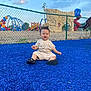toddler, child, playground, blue_surface, fence, outdoor, smiling, clothing, shoes, sky, clouds, play_equipment, slide, rock_climbing_wall, grass, tree, happy, person, daylight, fun