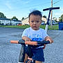 toddler, child, tricycle, outdoor, suburb, handlebars, clothing, shorts, tshirt, footwear, grass, sky, residential, road, toy, person, young_child, expression, daytime, vehicle