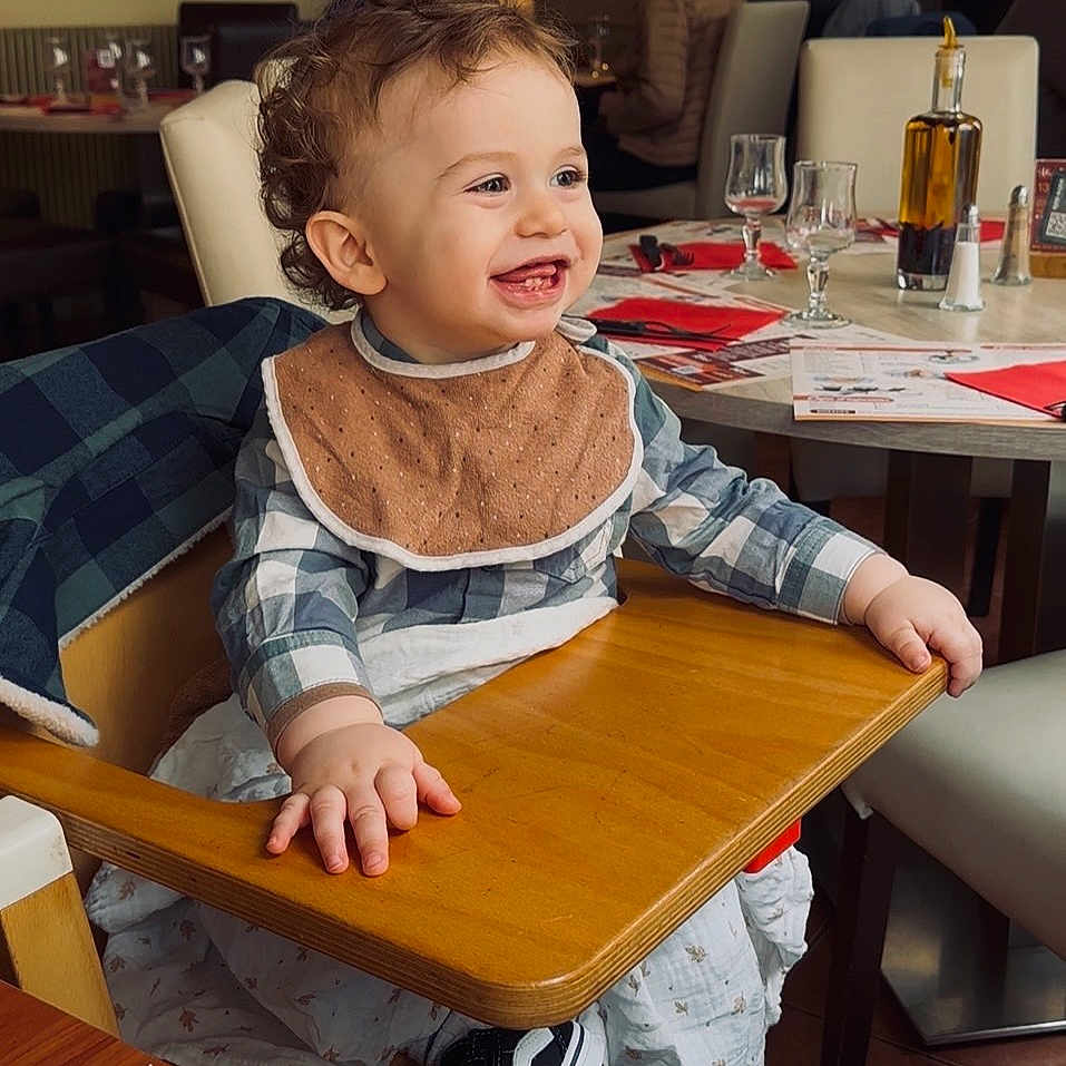 élio participe au concours pour gagner de l'argent avec cette photo : toddler, high_chair, smiling, indoor, restaurant, bib, curly_hair, table, chair, plaid_shirt, child, happy, wooden_surface, footwear, people_in_background, glassware, warm_lighting, decor, dining, casual_clothing