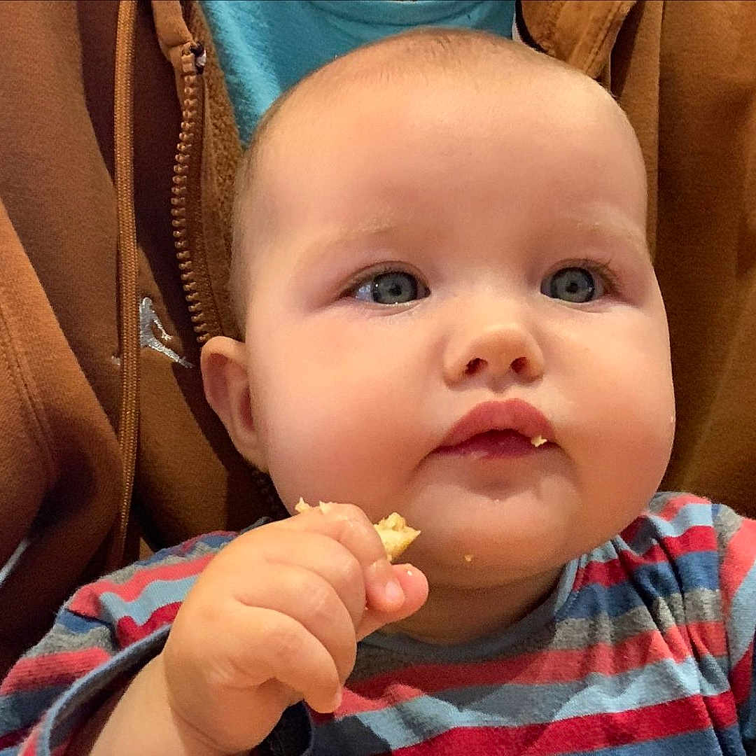 Ava a rejoint le concours — aidez-le/la à gagner de superbes lots ! baby, blue_eyes, brown_clothing, cheeks, child, closeup, clothing, cute, eating, face, food, hand, hoodie, indoor, munching, person, portrait, snack, striped_shirt, young_child