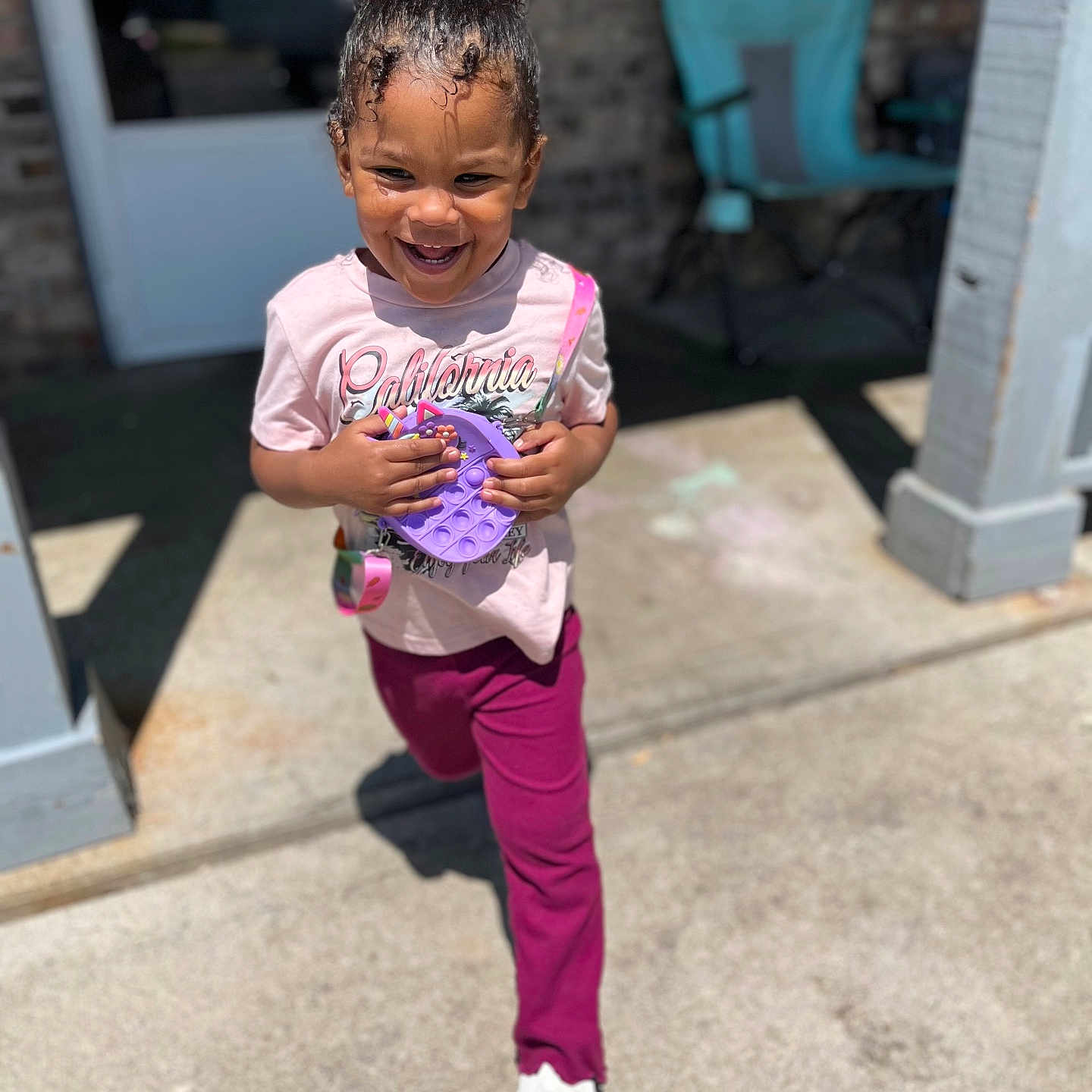 Alonna joined the competition — help win amazing prizes! brick_wall, bun, casual_clothing, chair, child, concrete, curly_hair, daylight, happy, outdoor, pants, playful, pop_it, porch, purple_toy, running, sandals, smiling, sunny, tshirt