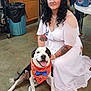 woman, dog, white_dress, tattoos, indoor, concrete_floor, bandana, bow_tie, black_and_white_dog, smiling_dog, curly_hair, pearl_necklace, trash_bin, table, stitch_bag, casual, kneeling, pet, person, happy