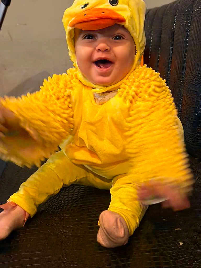 Khloe is registered to the contest to win money with this photo: baby, child, duck_costume, yellow, smiling, happy, cute, costume, plush, sitting, indoors, face, headwear, funny, joyful, hands, feet, infant, warm_clothing, playful