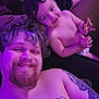 man, child, toddler, smile, tattoo, cookie, couch, curly_hair, beard, shirtless, indoor, purple_lighting, relaxing, family, happy, person, diaper, hand, face, living_room