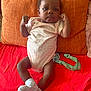 Katara is registered to the contest to win money with this photo: baby, infant, child, couch, pillow, orange_pillow, red_blanket, socks, onesie, heart_pattern, pacifier_clip, face, arm, leg, hand, furniture, indoor, cute, portrait, person