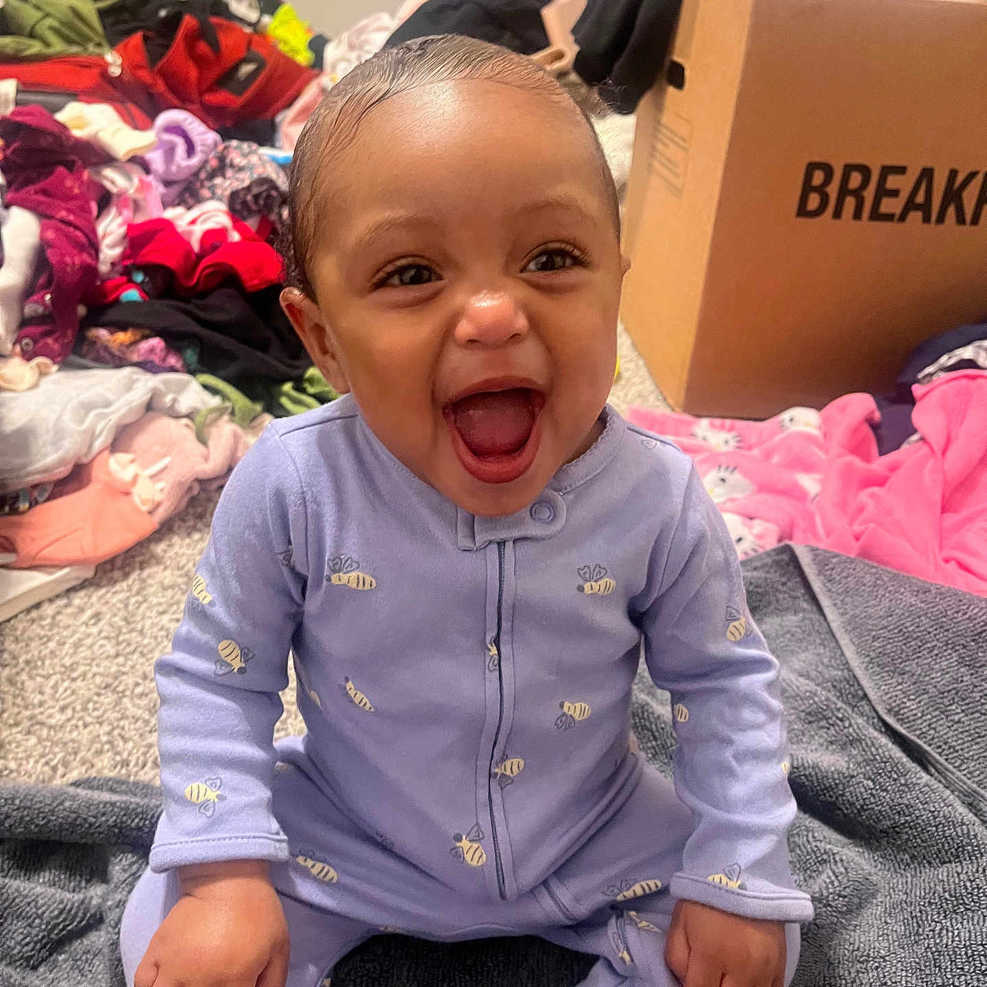 Aniyla is registered to the contest to win money with this photo: baby, bee_pattern, blanket, cardboard_box, carpet, child, clothes_pile, cute, face, hand, happy, head, indoor, infant, laughing, onesie, person, purple_clothing, sitting, smiling