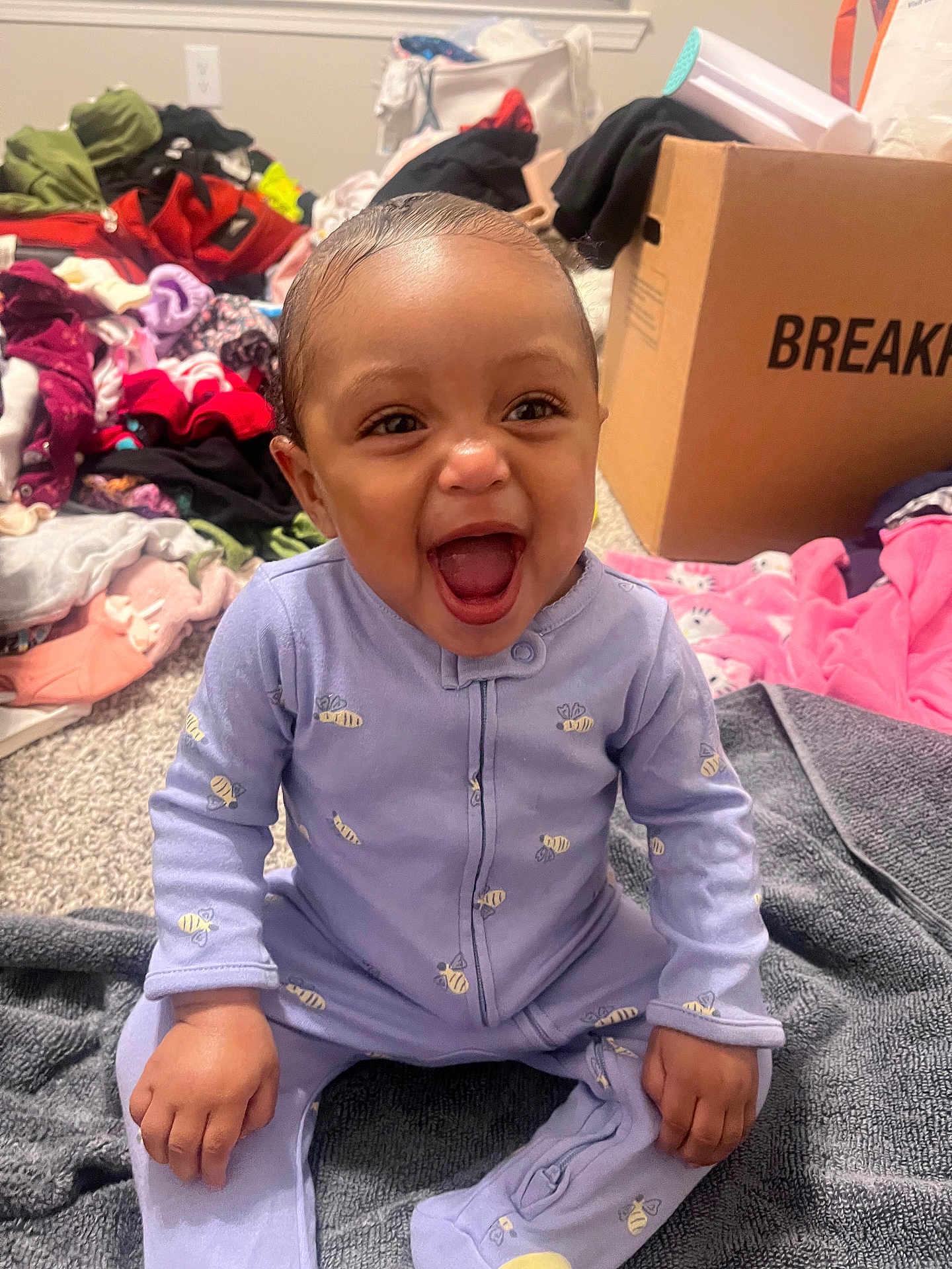 Aniyla is registered to the contest to win money with this photo: baby, child, smiling, laughing, onesie, purple_clothing, bee_pattern, indoor, carpet, blanket, clothes_pile, cardboard_box, happy, infant, person, cute, sitting, face, hand, head