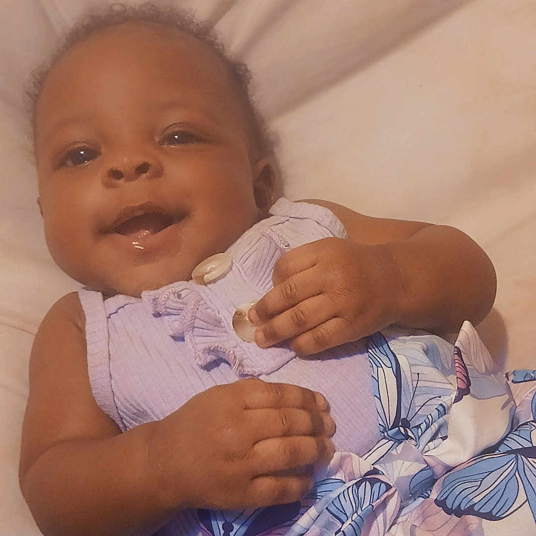 Dasia is registered to the contest to win money with this photo: baby, child, smile, purple_clothing, butterfly_pattern, dress, infant, lying_down, cute, happy, face, hand, soft_surface, indoors, emoji, heart, playful, young_child, portrait, skin